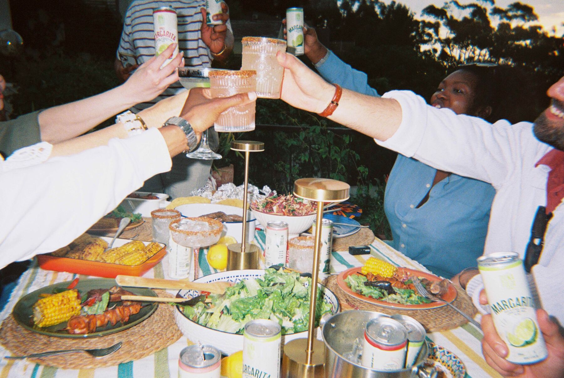 Friends toasting with Doña Fuego cans over a braai table of corn, salads and meat, shot on a disposable camera