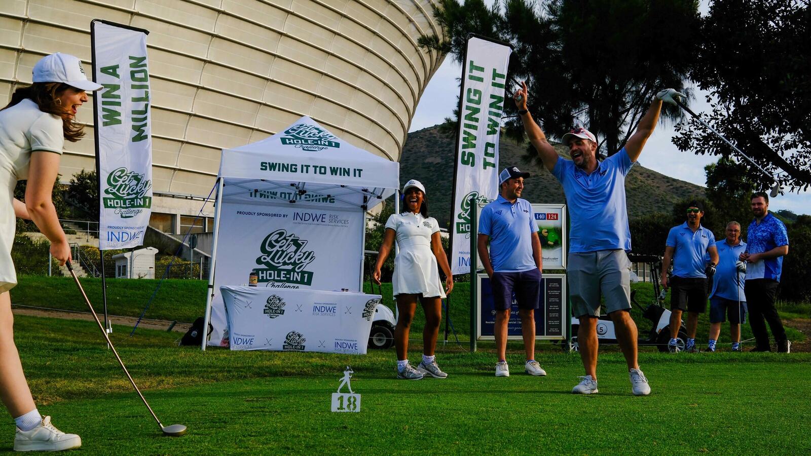 A golfer celebrating on a Get Lucky Hole-in-One Challenge tee at Cape Town Stadium, with Win a Million flags and an Indwe-branded gazebo